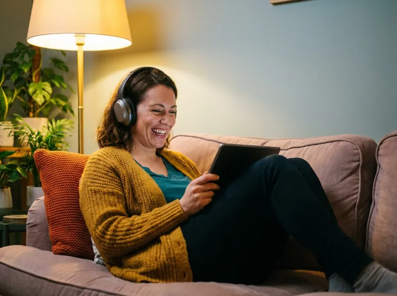 Woman with headphones smiling on a sofa while listening to an English audio story on a tablet